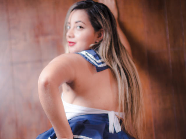 AbrilClark mit langem, blondem Haar, in einem blau-weißen Segleroutfit, mit dem Rücken zur Kamera, lächelnd, wobei ihr nackter Hintern und ein weißer Träger zu sehen sind, Holz im Hintergrund, weiches Licht..