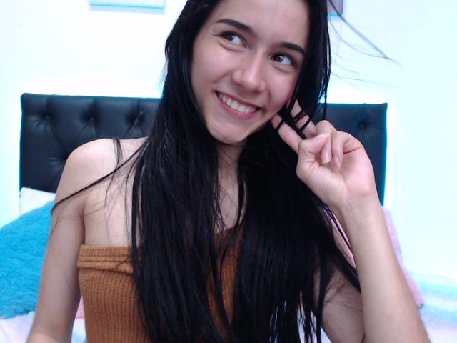 DeisyWest with long black hair, smiling, brown strapless top, showing her tits, hand near her face, blue pillow in background, black tufted headboard, white walls.