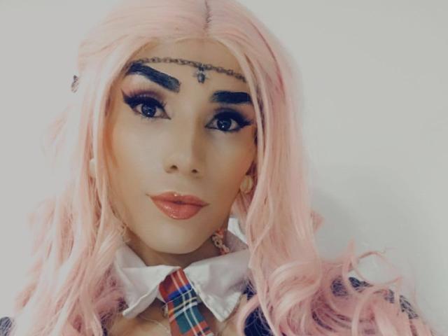 CrazyManHot has pink hair, bold eyebrows, and a forehead chain. They're wearing a plaid shirt with a white collar, and their makeup is dramatic with dark eyes.