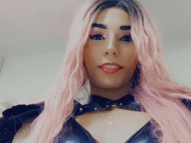 CrazyManHot with pink wavy hair, bold eyebrows, red lipstick, black leather choker, and black leather top, showing a cute smile and cute tits.