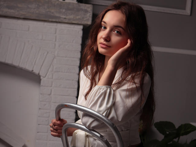 LuiMartin with long brown hair, white dress, sitting on metal chair, hand on cheek, soft smile, gentle light, white brick wall in background, subtle shadow, relaxed, cute.
