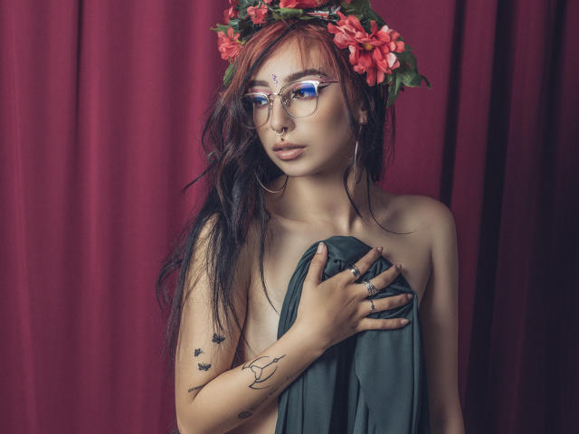 ConnieLuna met rood haar, een bloemenkrans, een bril, een neusring, oorringen, tatoeages, die groen materiaal vasthoudt, haar borsten bedekkend, voor een rode gordijn, kijkend naar de camera, met een zachte uitdrukking..