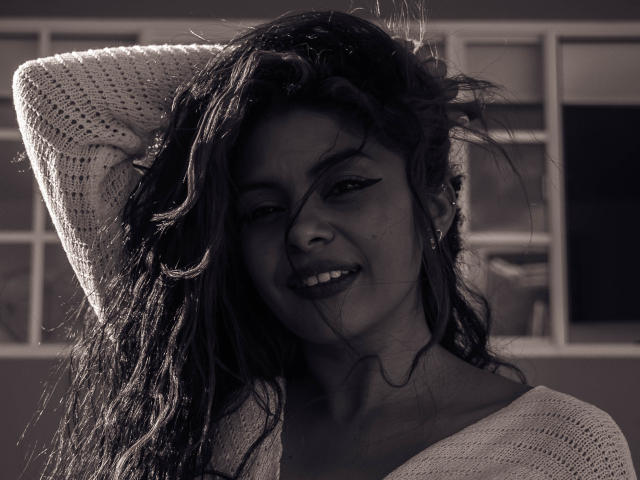 Anna, met golvend donker haar, lachend, hand in trui, schaduw op het gezicht, zacht licht, zwart-wit, iets rommelig haar, subtiele make-up, trui iets bloot aan de schouders, schattig..