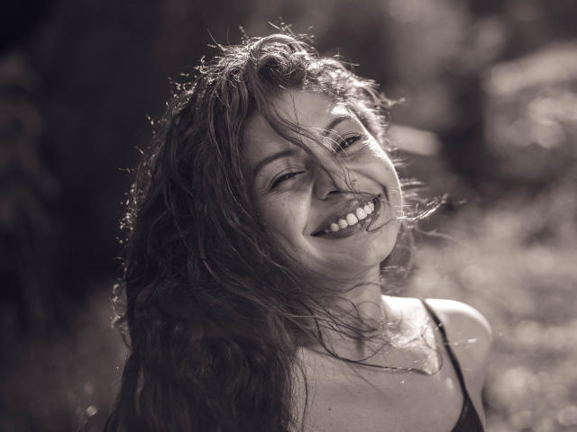 AnnaMesht's face, smiling with her eyes closed, has curly wet hair in her face, showing her teeth. She's wearing a black spaghetti strap top, and sunlight highlights her.