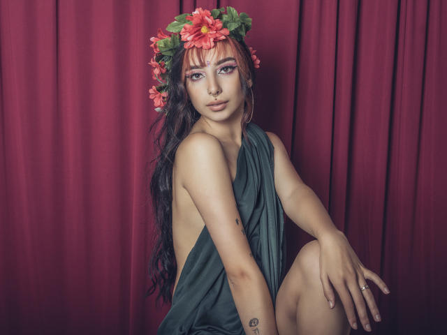 ConnieLuna met lang zwart haar, roze bloemen in haar hoofd, een groene jurk, met een open rug, zittend, een rode gordijn, een tatoeage op haar arm, een neusring, een ring aan haar vinger, kijkend naar de camera..
