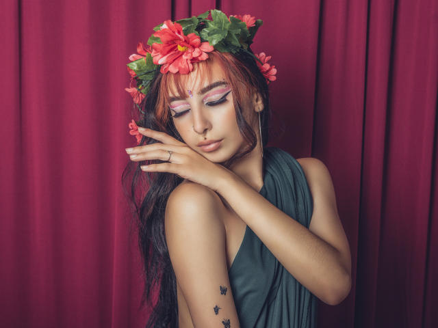 ConnieLuna, com cabelos longos e pretos, sombra rosa e roxa, coroa de flores, vestido verde, tocando o rosto, tatuagem de estrelas pretas no braço direito, fundo com cortina vermelha..