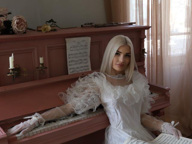 PleasingAngel with platinum blonde hair, wearing a white, ruffled, Victorian-style dress and gloves, sitting at a pink piano, holding sheet music, with candles on the piano.