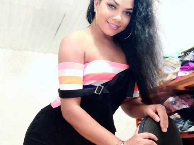 AbellaPhill with long curly black hair, pink-striped off-shoulder top, black dress, and hoop earrings, smiling at camera, showing her titties and cute pink lipstick.