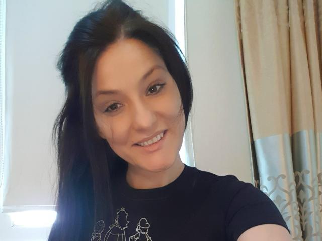 JoliDanielle with long black hair, smiling, wearing a black shirt with white tree designs, showing her cute face and small nose, standing in front of a window.