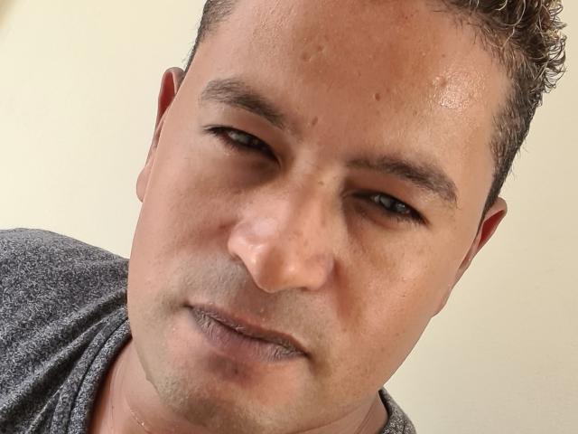 MrBlackHot hat ein verschwitztes, leicht lockiges Gesicht mit einem neutralen Ausdruck und trägt ein graues Hemd. Seine Nase ist klein und seine Haut ist hellbraun mit sichtbaren Poren..