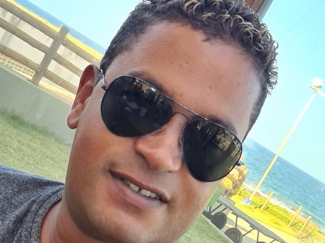 MrBlackHot ha i capelli castano ricci, indossa occhiali da sole neri da aviatore e una camicia grigia, con un viso leggermente sudatore e un piccolo sorriso, ed è in piedi vicino all'oceano..