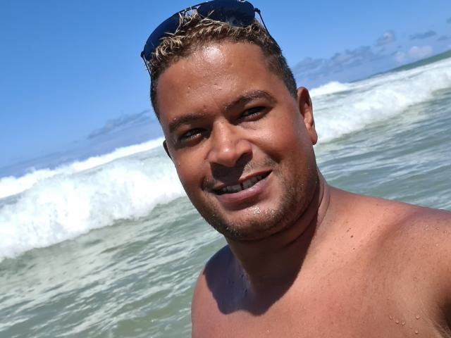 MrBlackHot zonder shirt, met kort, blond haar, een lichte baard, en een blauwe lucht op de achtergrond, lacht naar de camera met een zonnebril op zijn hoofd, en staat in de oceaan met golven..