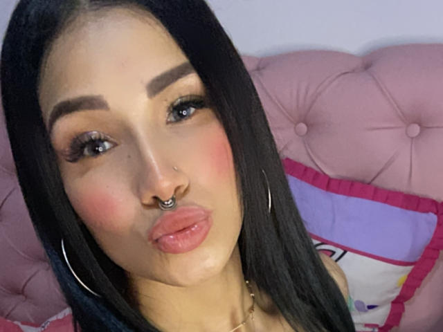 OnniOconor with black hair, hoop earrings, nose ring, pouty pink lips, and bold eyebrows, wearing makeup, puffed cheeks, sitting on pink tufted bed.