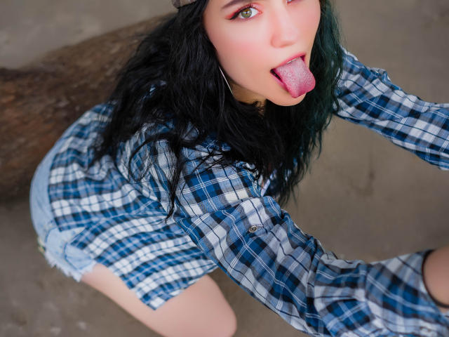 MolySmith, com olhos verdes, cabelo preto ondulado e língua rosa para fora, vestindo uma camisa xadrez azul e shorts de jeans, ajoelhada em um piso de concreto, mostrando o bumbum..