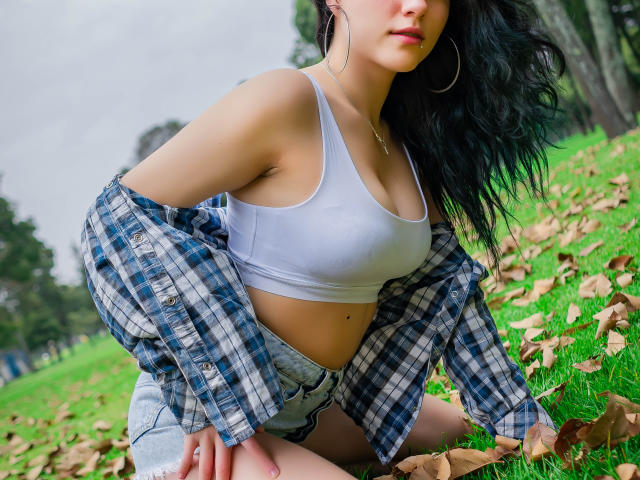 MolySmith, agenouillée sur l'herbe, portant un débardeur de sport blanc qui révèle ses petites seins, et une chemise à carreaux bleus enroulée autour de ses épaules, avec un short en jean et des boucles d'oreilles cercles..