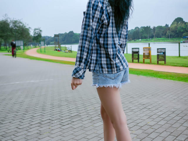 MolySmith trägt ein blau-weiß kariertes Hemd und zerrissene Jeansshorts. Sie steht mit dem Rücken zur Kamera und zeigt ihr Gesäß auf einem befestigten Weg..