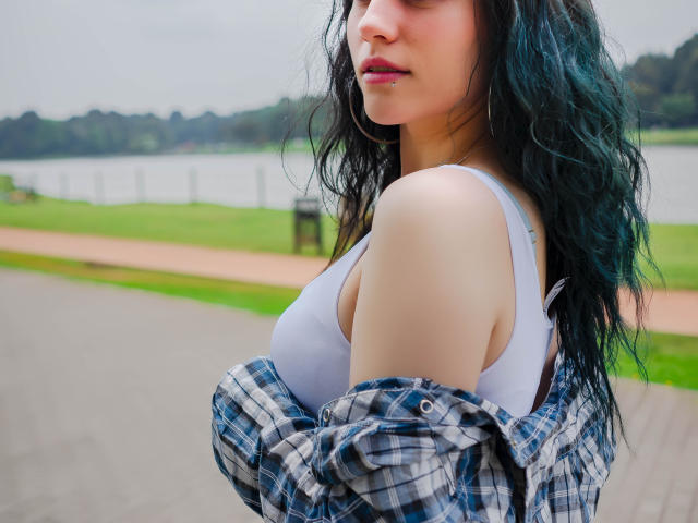 MolySmith, con i capelli ondulati color turchese, reggiseno sportivo bianco e camicia a quadri blu che lascia scoperte le spalle, mostrando il seno, in piedi vicino a un fiume, che guarda di lato, con un'espressione accigliata..