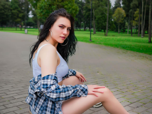 MolySmith, con i capelli neri e ondulati, indossa una canotta bianca, una camicia a quadri e orecchini a cerchio, seduta su un vialetto di mattoni, mostrando la biancheria intima bianca e il sedere scoperto..