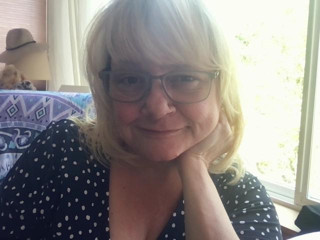 ItsAllMine mit blonden Haaren, Brille und einem leichten Lächeln, mit dem Kinn in der Hand, trägt ein schwarzes Top mit Punkten und sitzt am Fenster..