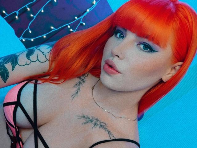 BrunnaCatie mit leuchtend orangefarbenem Haar, blauen Augen und schwarzem Eyeliner, die ihre großen Brüste in einem pinken und schwarzen Top mit Trägern zeigt, mit Tattoos an Arm und Brust und einem Nasenring..