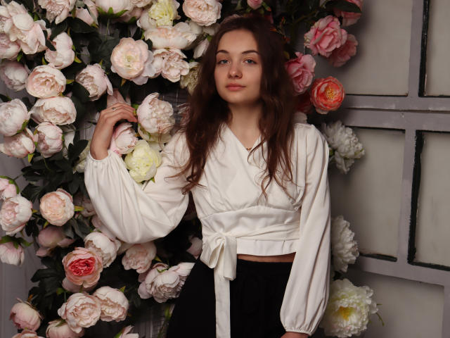 LuiMartin with wavy brown hair, wearing a white blouse tied at the waist, black pants, standing among pink and white flowers, gently touching her hair.