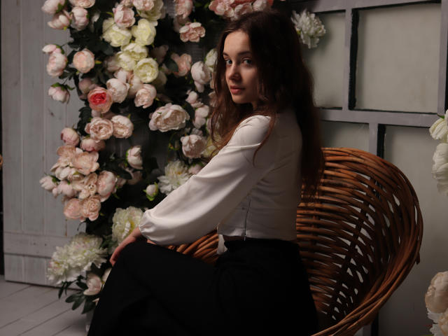 LuiMartin with wavy brown hair, white blouse, black skirt, sitting on a wicker chair, surrounded by pink and white roses, looking over her shoulder with a slight smile.
