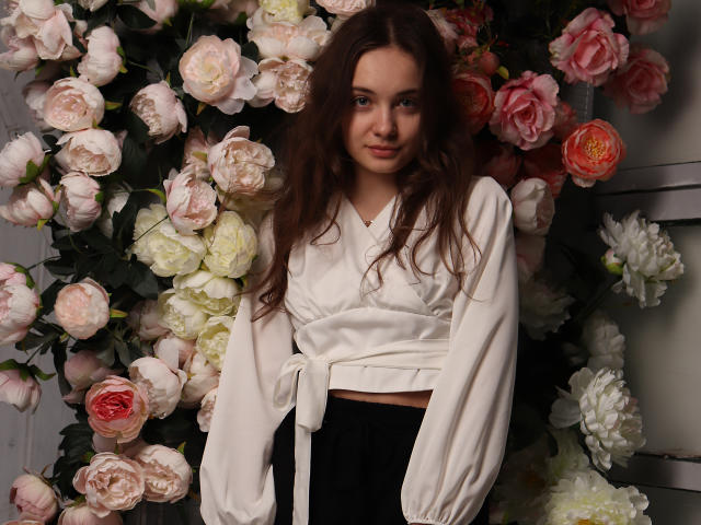 LuiMartin, con lunghi capelli castani, indossa una maglia bianca a maniche lunghe, annodata, pantaloni neri e si trova di fronte a un muro fiorito con rose rosa, bianche e rosse..
