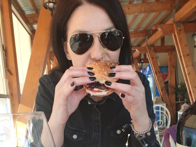 JoliDanielle with black hair, wearing sunglasses and a denim shirt, eats a burger with both hands, black nail polish, and a glass on the table in front.