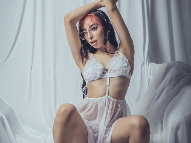 ConnieLuna mit rosa und schwarzen Haaren, trägt einen weißen Spitzen-BH und ein durchsichtiges Babydoll, die Arme sind erhoben, sie sitzt mit gespreizten Beinen und zeigt ihre Brüste und Vulva durch den transparenten Stoff..