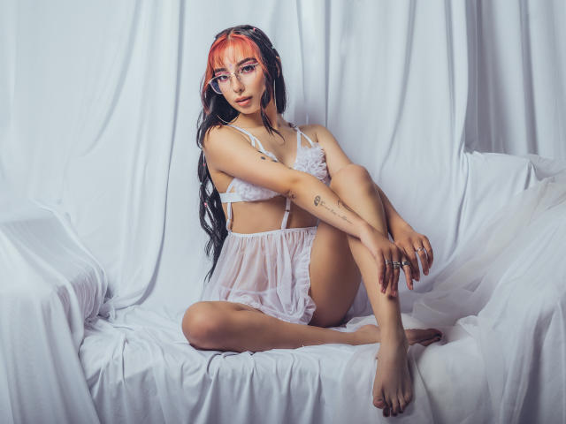 ConnieLuna mit rot und schwarz gefärbtem Haar, die zarte weiße Dessous trägt und auf einem weißen Stoff sitzt, die Beine gekreuzt hat, wobei Tattoos und ein Ring an ihrem Finger sichtbar sind..