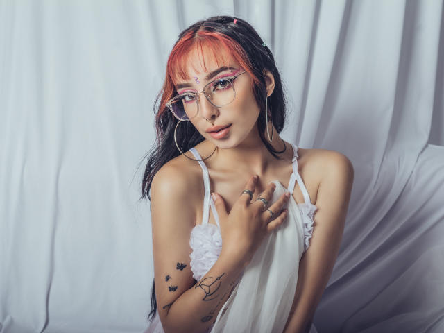 ConnieLuna con capelli arancioni e neri, occhiali, vestito bianco, tatuaggi, anelli, che tiene un vestito, guarda la fotocamera, sfondo bianco drappeggiato. Seno visibile, carina, attraente, femminile..