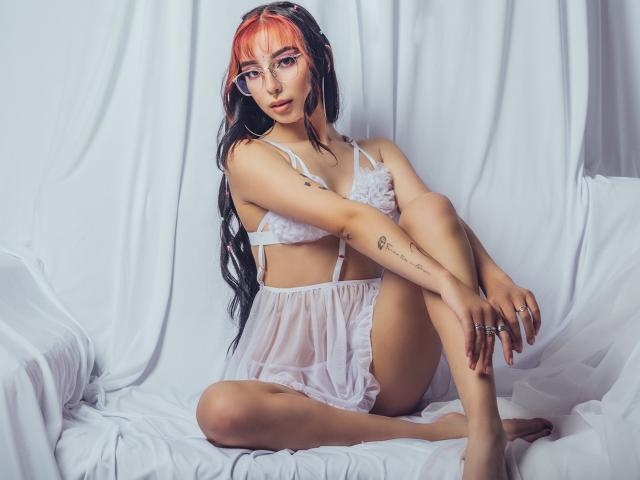 ConnieLuna mit orange-schwarzem Haar, Brille, weißer Unterwäsche, sitzend auf einem weißen Bett, Beine gekreuzt, Armtätowierungen, blickt in die Kamera und zeigt ihre Brüste und Vulva durch einen durchsichtigen Stoff..