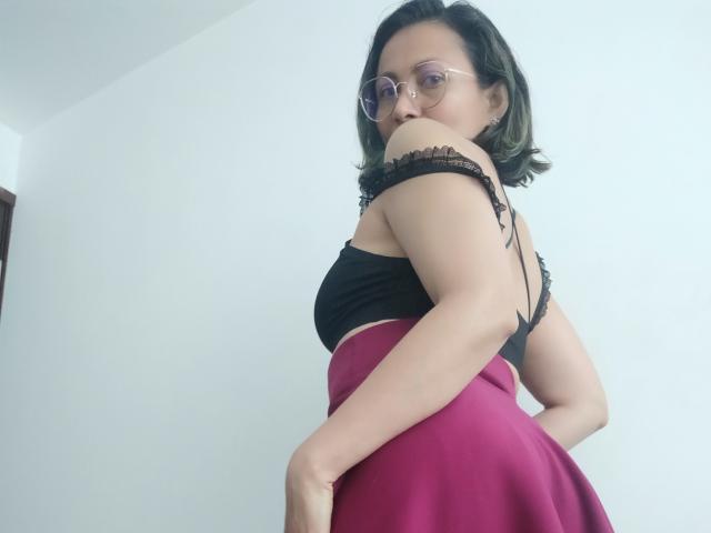 AlishaColins con gli occhiali, una canottiera nera con spalline, e una gonna rosa, che mostra la schiena e i glutei, una parte in pizzo nero a spalle scoperte, capelli castani corti, che guarda oltre la spalla..