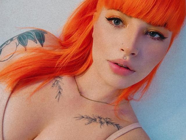 Brunna Catie, com cabelo laranja vibrante, olhos azuis e tatuagens no ombro e no peito, vestindo uma blusa rosa, usa um piercing no nariz e um piercing no lábio..
