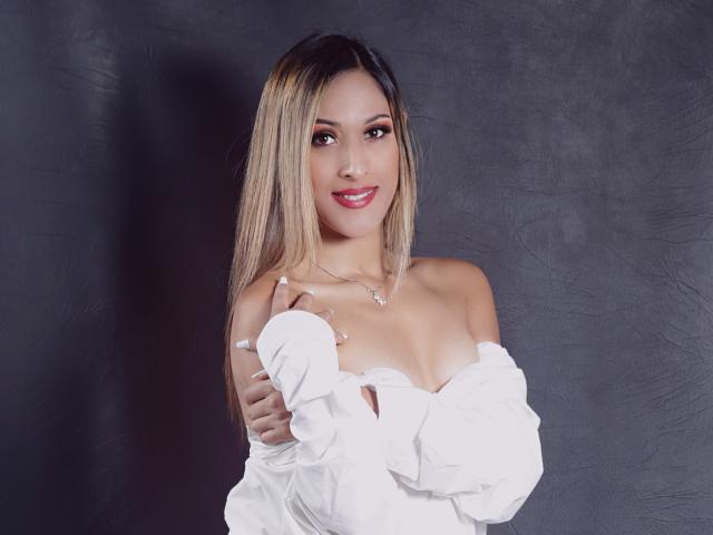 EstrellaHarryson with blonde hair, smiling, wearing a white off-shoulder top, exposing her tits, holding the top down, grey background, red lipstick, white nails, necklace.