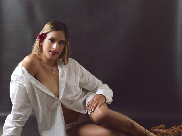 EstrellaHarryson with blonde hair, red flower in her hair, wearing an open white shirt, showing her tits, and white lace panties, sitting with one knee up.