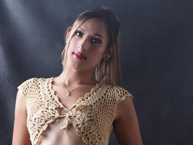EstrellaHarryson with tan skin, dark hair, red lipstick, wearing a beige crocheted top that barely covers her small tits, looking at the camera with a slight smile.