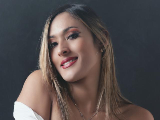 EstrellaHarryson has blonde hair, red lipstick, and is smiling with her head tilted. She's wearing a white off-shoulder top, and her tits are partially visible.