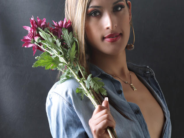EstrellaHarryson with blonde hair, wearing a denim shirt, holding pink flowers, gold hoop earrings, and a necklace, looking at the camera with a slight smile.