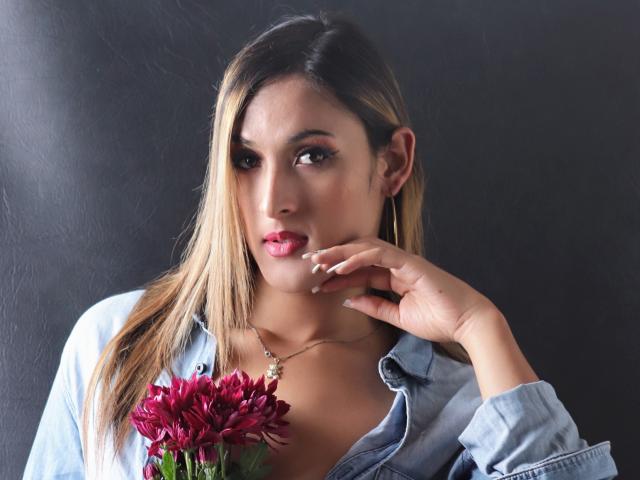 EstrellaHarryson with blonde hair, wearing a blue shirt, holding a red flower, looking serious, hand near mouth, dark background, necklace with small pendant, cute and sexy.