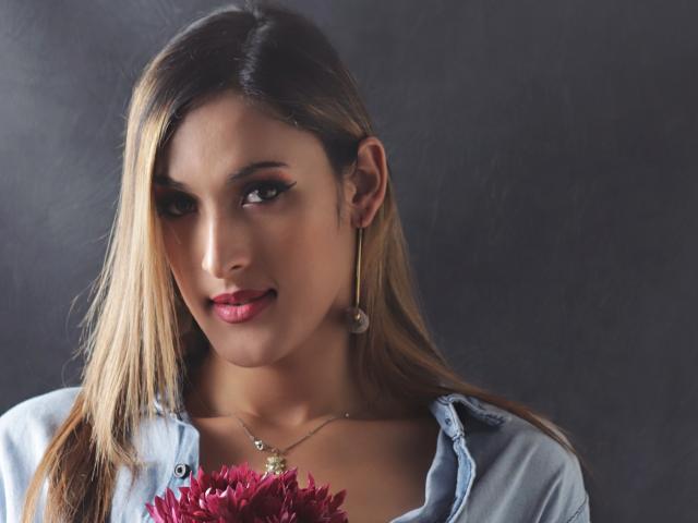 EstrellaHarryson has blonde hair, dark eyebrows, and red lipstick, wearing a denim shirt, holding red flowers, with a subtle smile, and a necklace with a small pendant.