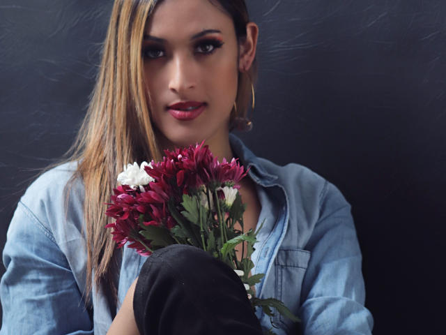 EstrellaHarryson with long blonde hair, blue denim shirt, holding purple and white flowers, black pants, hoop earrings, dark background, serious expression, pink lipstick, sexy.