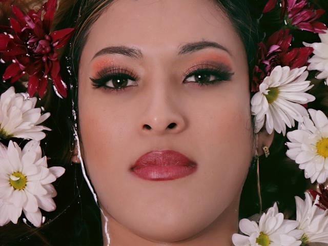 EstrellaHarryson's face with red lipstick, pink eyeshadow, and black eyeliner, surrounded by white daisies and red flowers, her brown hair partly visible.