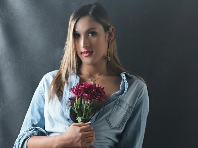 EstrellaHarryson with long blonde hair, blue denim shirt, holding red flowers, gold hoop earrings, and red lipstick, looking slightly to the side with a subtle smile.