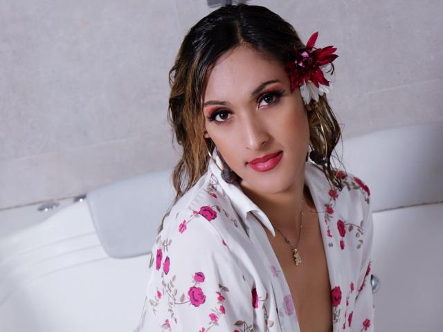 EstrellaHarryson has wavy brown hair with a red and white flower, wearing a floral shirt, pink lipstick, and a necklace, sitting in a white-tiled bathroom.