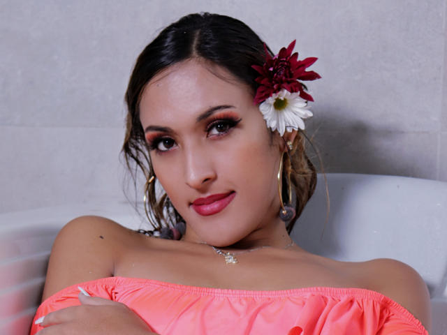 EstrellaHarryson with dark hair, pink off-shoulder top, red and white flower in hair, hoop earrings, and pink lipstick, arms crossed, looking at camera, smiling, sitting in white bathtub.