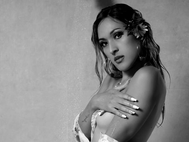 EstrellaHarryson with wet hair, a flower in her hair, and her hands covering her tits, water spraying her. She has long nails, a tattoo on her arm, and hoop earrings.