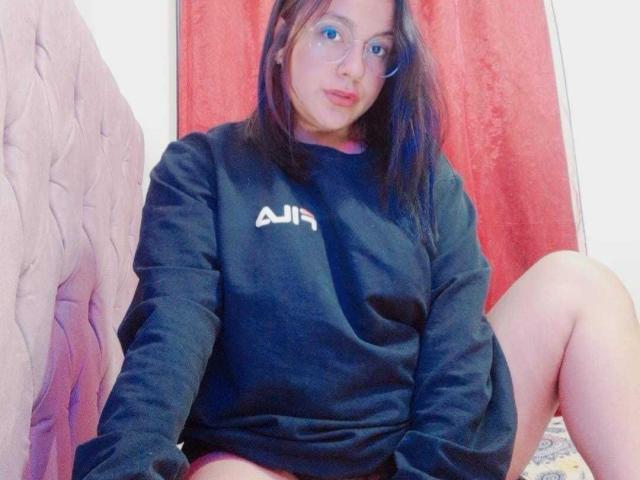EmmaLuna, avec ses cheveux bruns, ses lunettes et son pull noir ASIIR, assise les jambes écartées, montrant ses cuisses, sur un lit rose, devant un rideau rouge..
