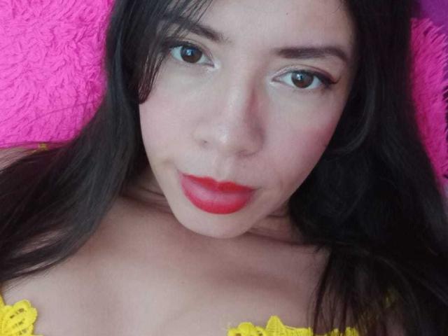 Une femme, AnyJhonsHentay, avec des lèvres rouges, des cheveux noirs et un haut en dentelle jaune, regardant l'appareil photo avec des yeux marrons, allongée sur une surface rose texturée. Les seins sont partiellement visibles..