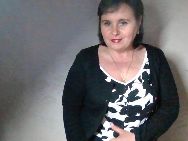VadaHot, avec ses cheveux noirs, son rouge à lèvres rouge, portant un cardigan noir par-dessus un haut blanc et noir à motifs floraux, et un collier, souriant à l'appareil photo. Ses mains touchent doucement sa poitrine..